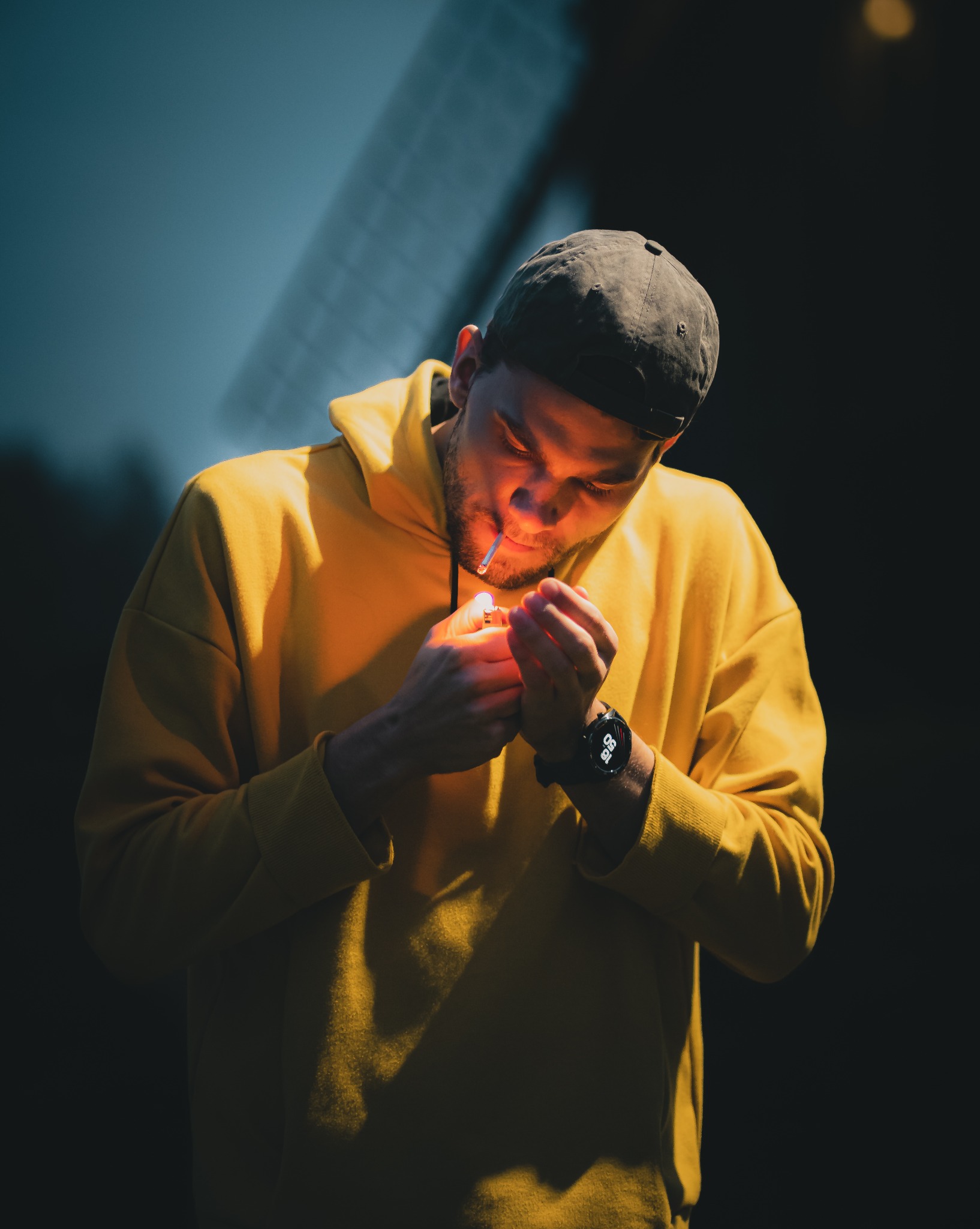 Mężczyzna w żółtej bluzie z kapturem zapala papierosa zapalniczką. Widoczny zegarek na ręce, ciemne tło i oświetlenie od zapalniczki.