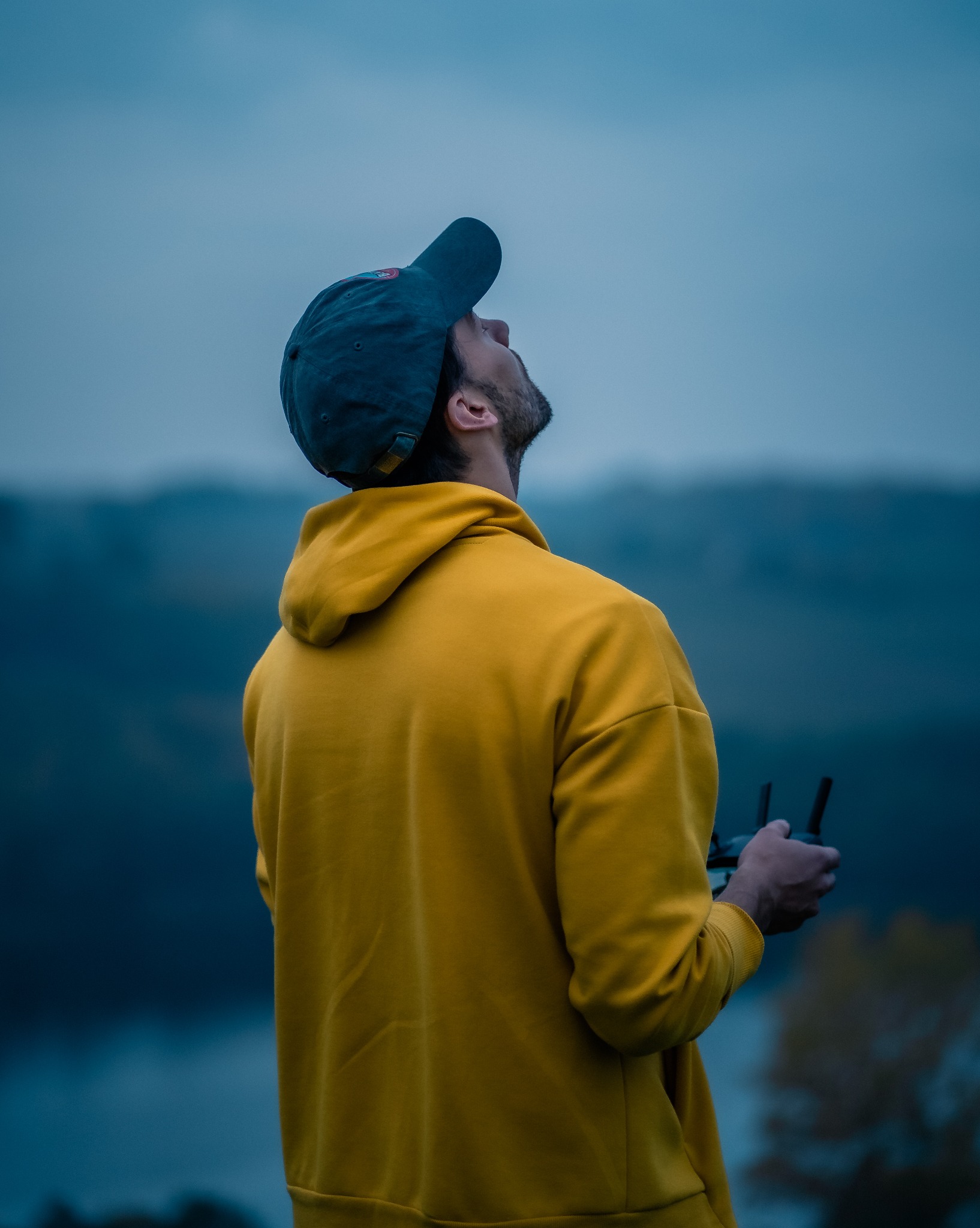 Mężczyzna w żółtej bluzie i czapce spogląda w górę, trzymając kontroler drona. W tle rozmyty krajobraz z jeziorem i drzewami w pochmurny dzień.