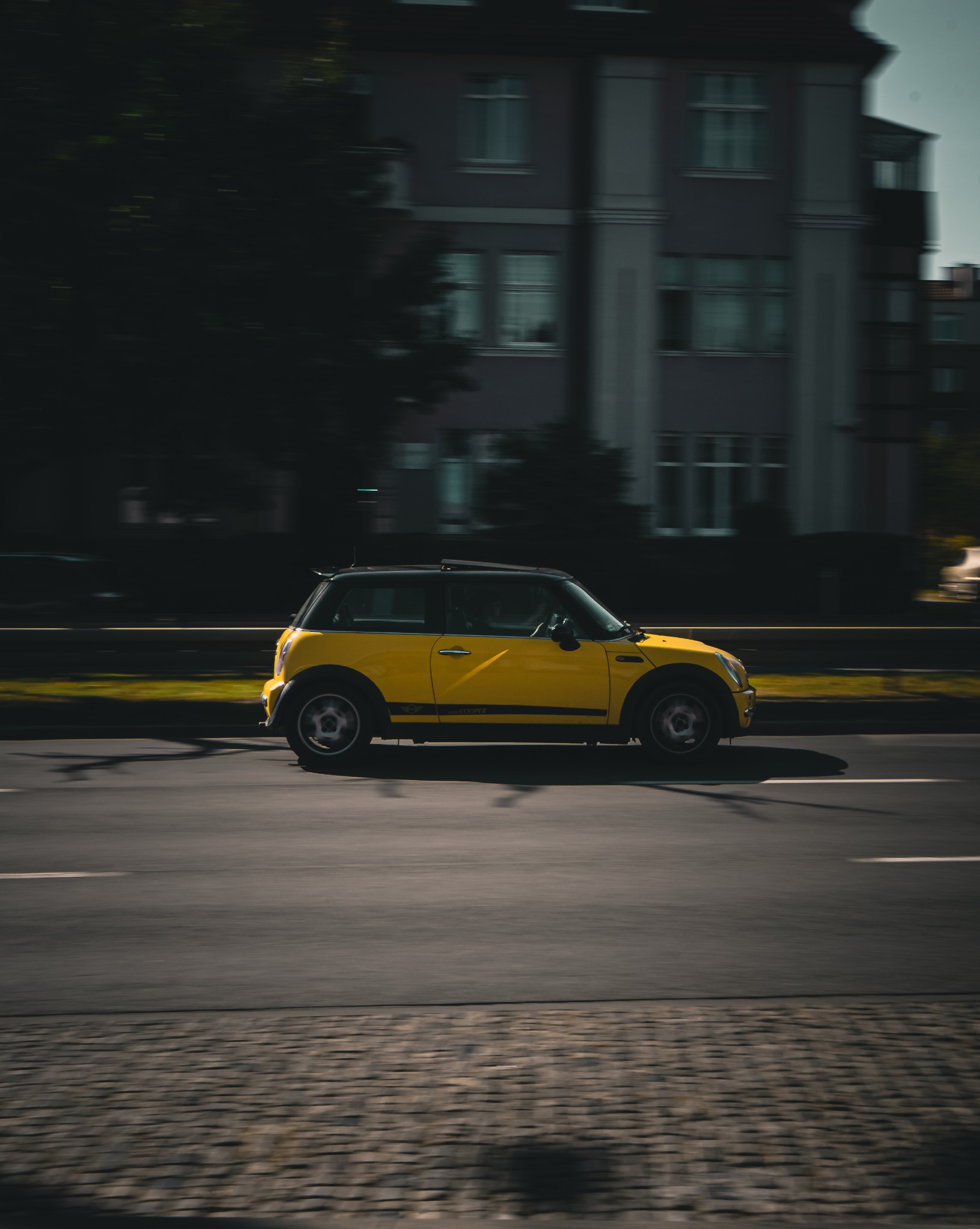 Żółty Mini Cooper w ruchu, uchwycony w dynamicznym ujęciu z rozmytym tłem miejskim, podkreślającym prędkość i energię. Czarny dach i felgi kontrastują z jaskrawym lakierem.