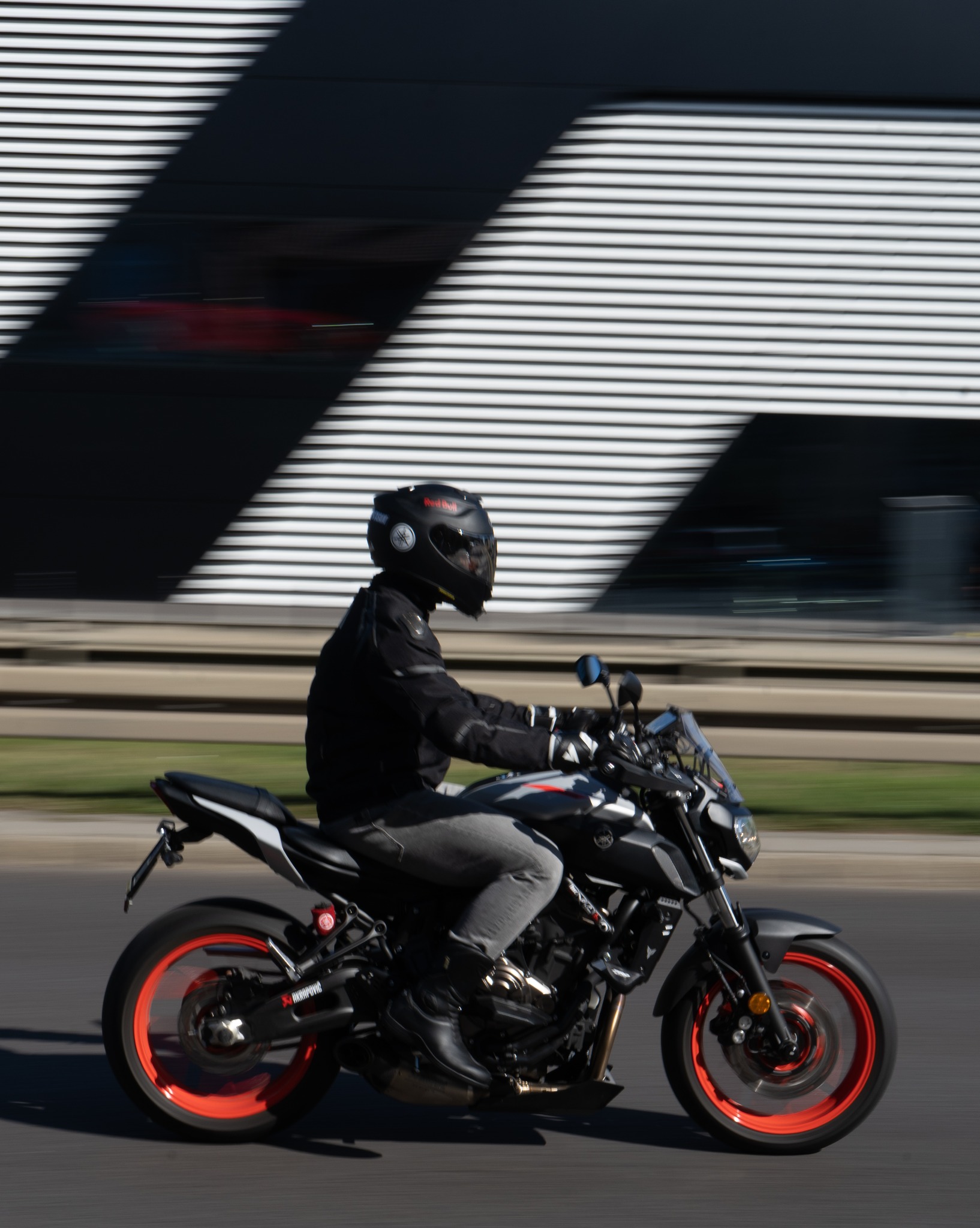 Dynamiczne ujęcie motocyklisty na czarnym motocyklu z czerwonymi felgami, na tle nowoczesnego budynku z poziomymi pasami. Ruch podkreślony rozmyciem tła.