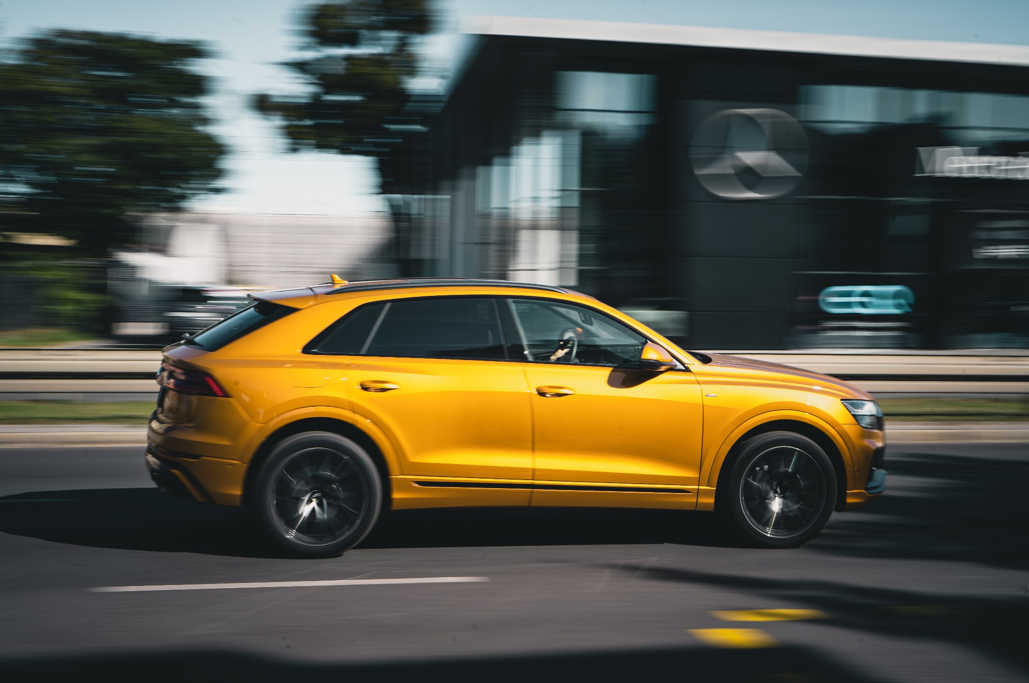 Dynamiczne ujęcie złotego Audi Q8 w ruchu, z rozmytym tłem miejskim i salonem Mercedesa w tle. Efektowna fotografia motoryzacyjna.