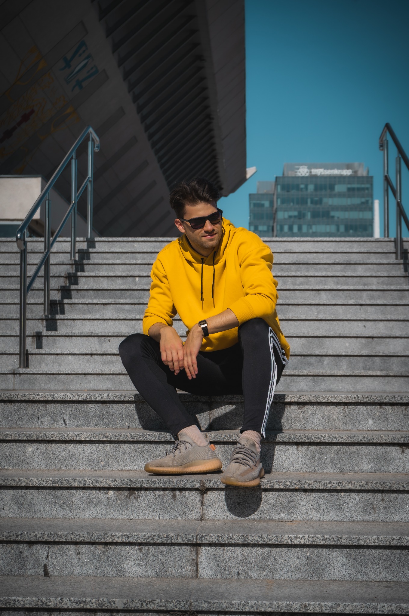 Mężczyzna w żółtej bluzie i okularach siedzi na schodach, w tle budynki biurowe i błękitne niebo. Ujęcie z dołu, podkreślające miejski charakter sesji.
