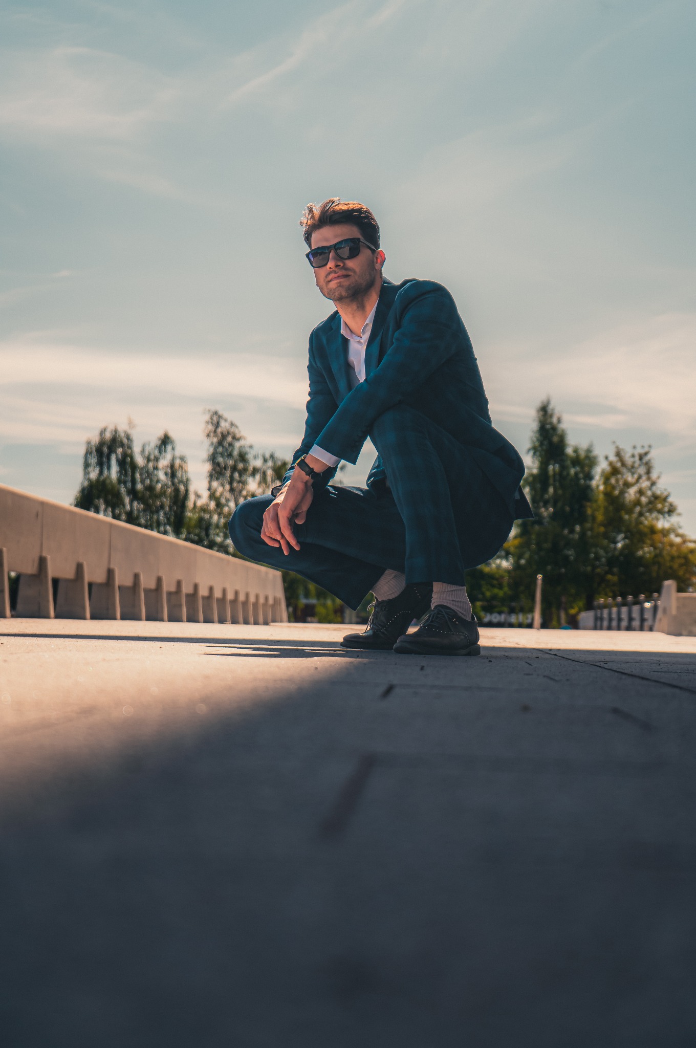 Mężczyzna w garniturze w kucki, z ciemnymi okularami, na tle miejskiego krajobrazu z betonową barierką. Ujęcie z dołu, w słoneczny dzień.