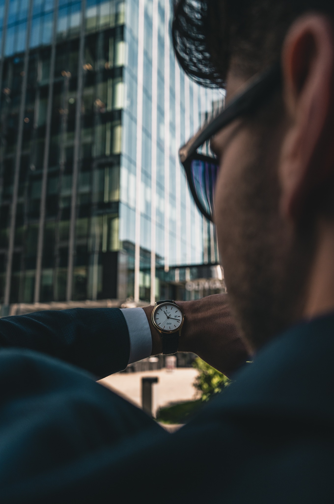 Mężczyzna w garniturze spogląda na zegarek na tle szklanego biurowca. Widok zza ramienia, ciemna tonacja, odbicia w okularach.