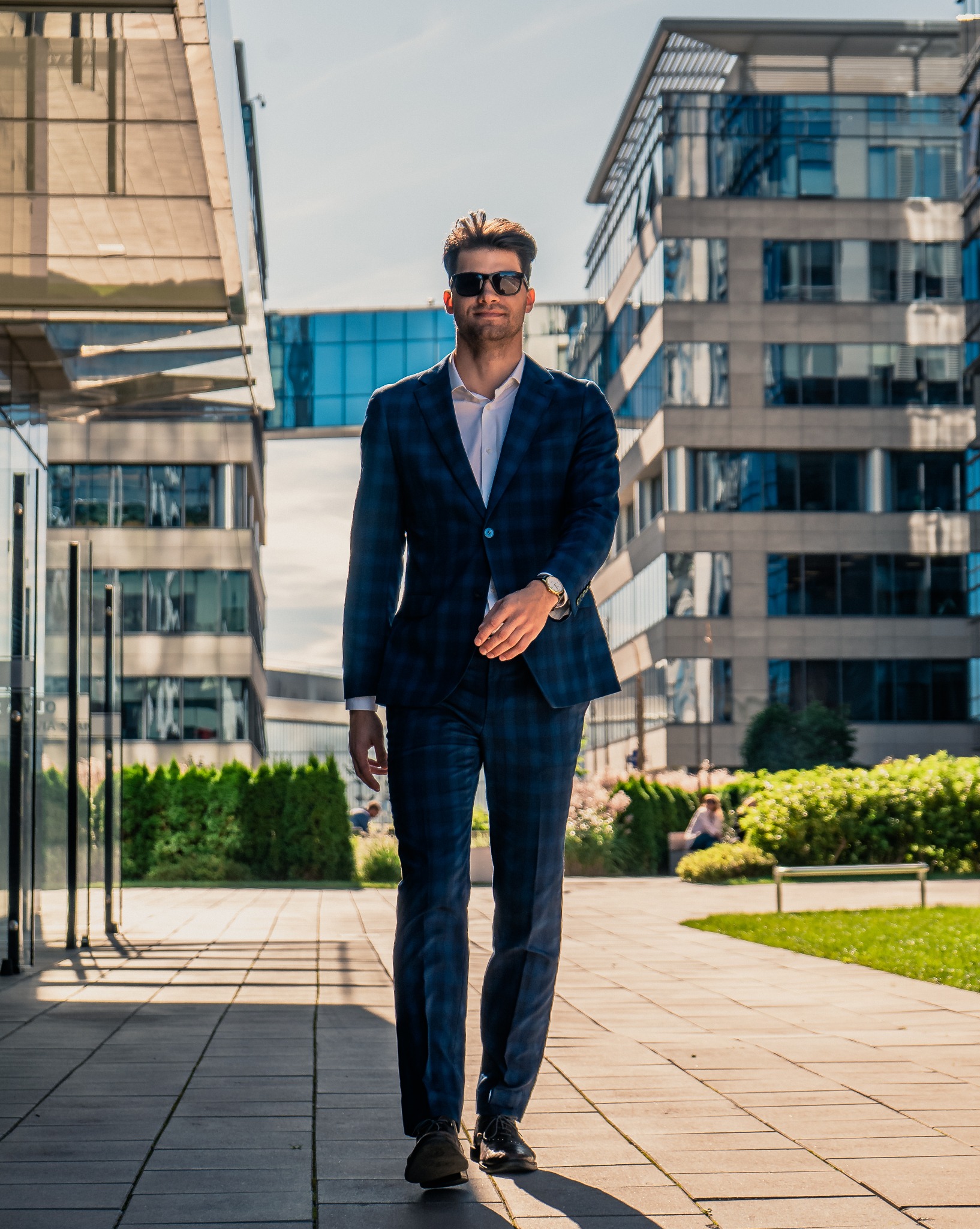 Mężczyzna w garniturze w kratę i okularach przeciwsłonecznych idzie chodnikiem na tle nowoczesnych budynków biurowych w słoneczny dzień. Dynamiczne ujęcie.