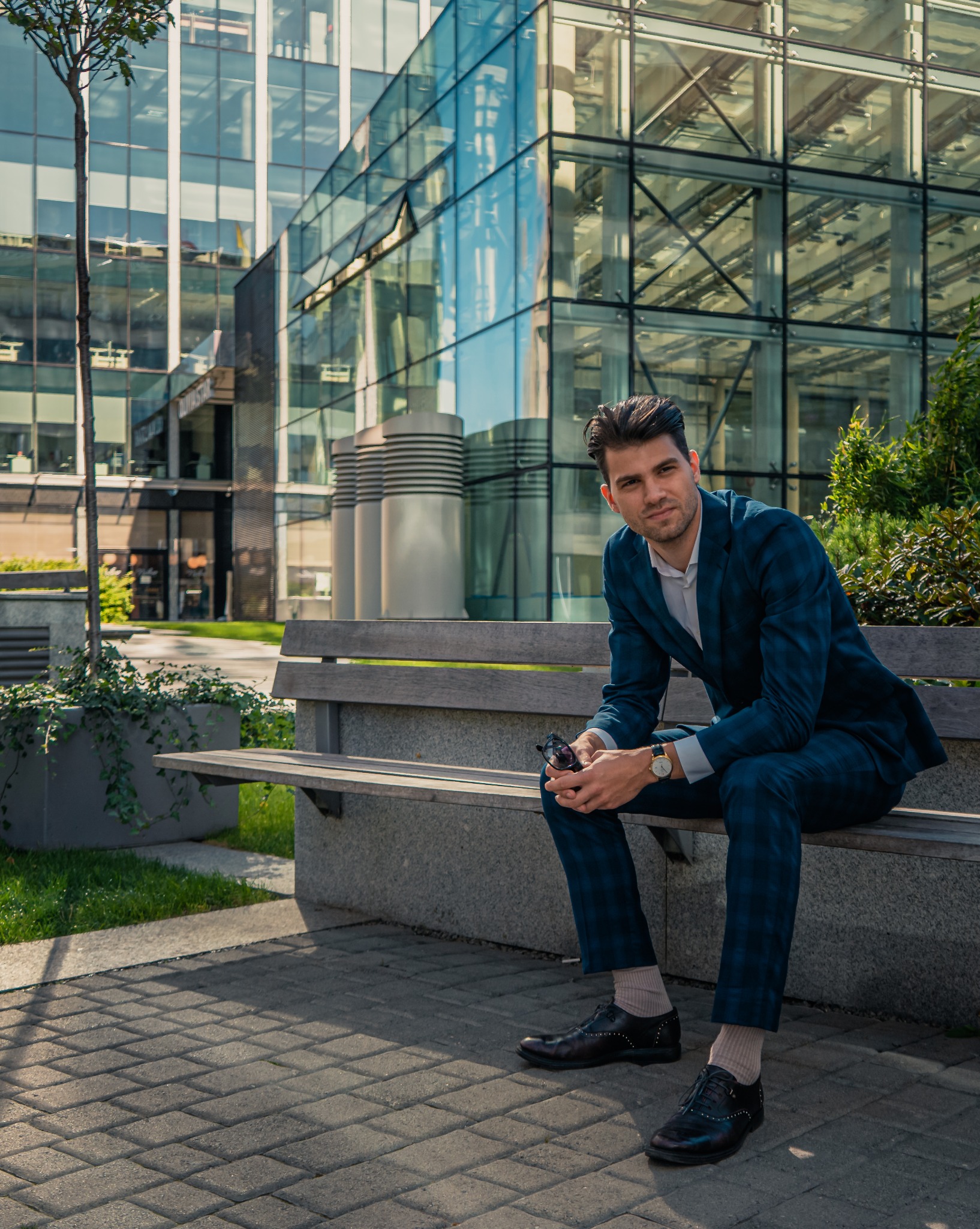 Mężczyzna w garniturze w kratę siedzi na ławce przed nowoczesnym biurowcem ze szkła. W ręku trzyma okulary, na nadgarstku zegarek. Elegancki styl i miejski krajobraz.
