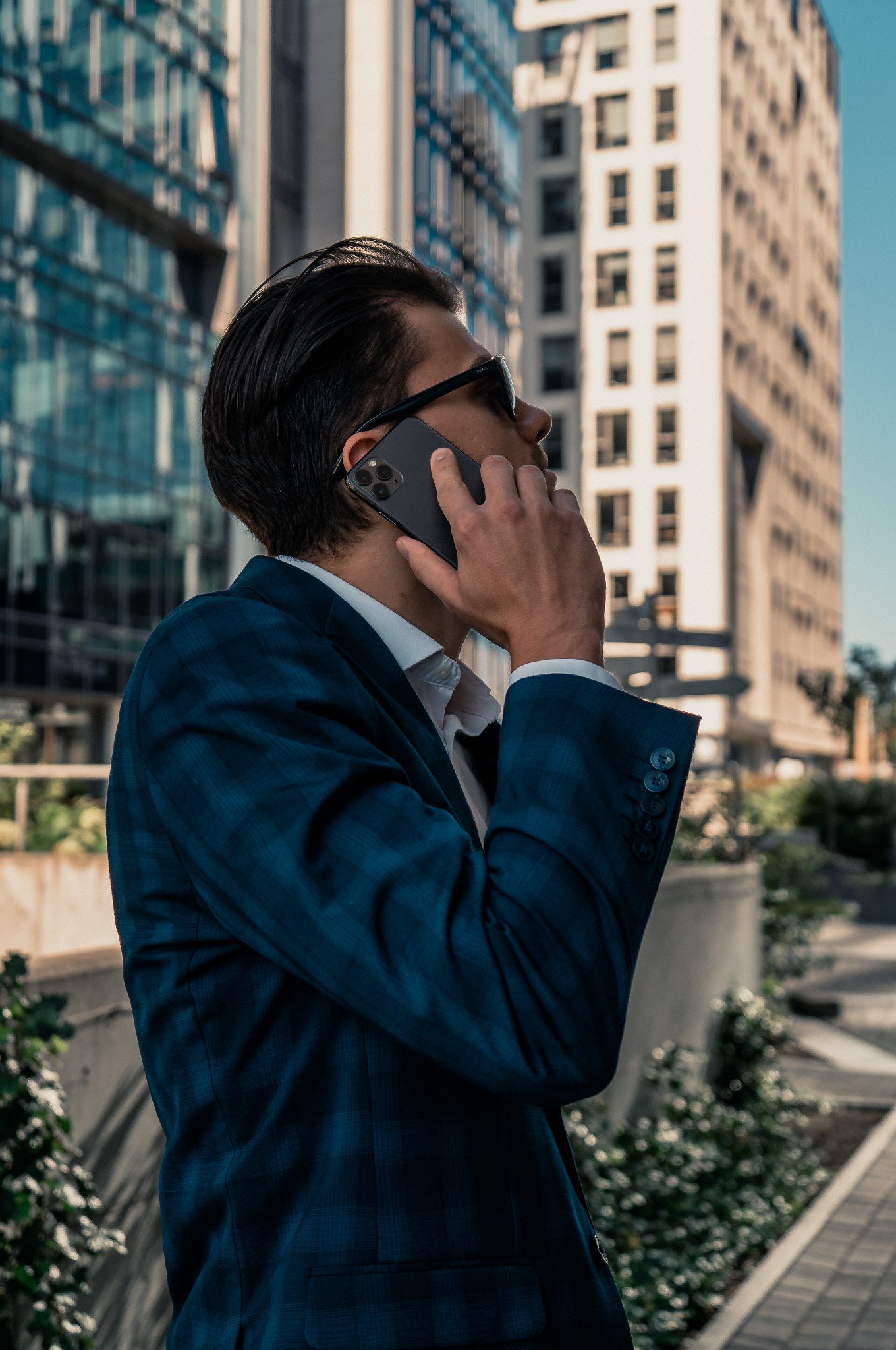 Mężczyzna w niebieskiej marynarce rozmawia przez telefon na tle nowoczesnych budynków. Widoczny profil twarzy i fragment sylwetki.