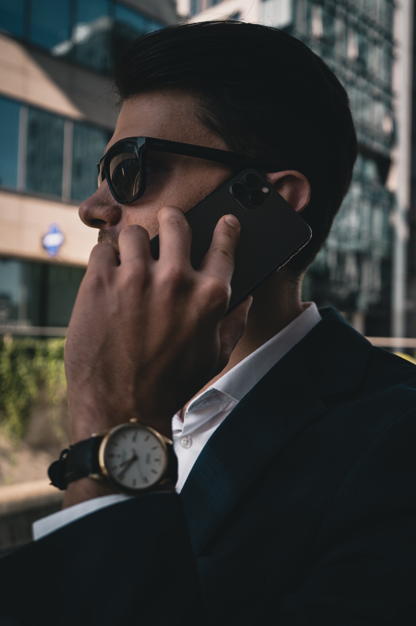 Mężczyzna w garniturze, z zegarkiem na ręku, rozmawia przez telefon na tle nowoczesnego budynku. Ujęcie z profilu, ciemne okulary.