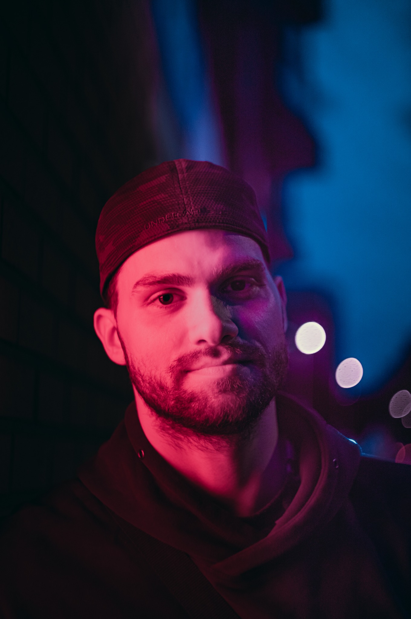 Portret mężczyzny w czapce z brodą, oświetlonego czerwonym i niebieskim światłem, z efektem bokeh w tle, tworzącym unikalną atmosferę. Nietypowe oświetlenie.