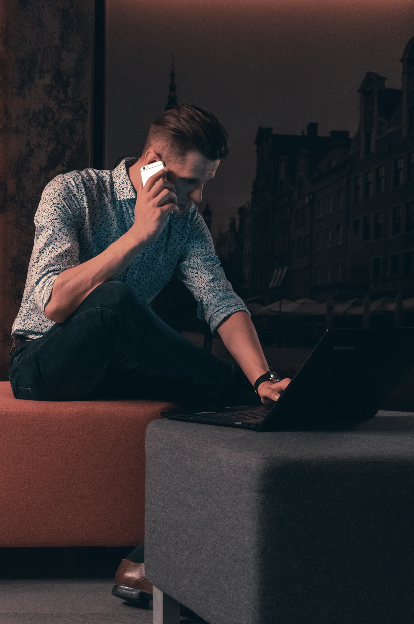 Młody mężczyzna w koszuli rozmawia przez telefon i pracuje na laptopie, siedząc na nowoczesnym siedzisku w biurze. W tle widoczny zarys panoramy miasta.