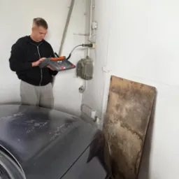 Elektryk sprawdza instalację elektryczną w garażu za pomocą miernika. Widoczne przewody, puszki elektryczne i fragment samochodu.