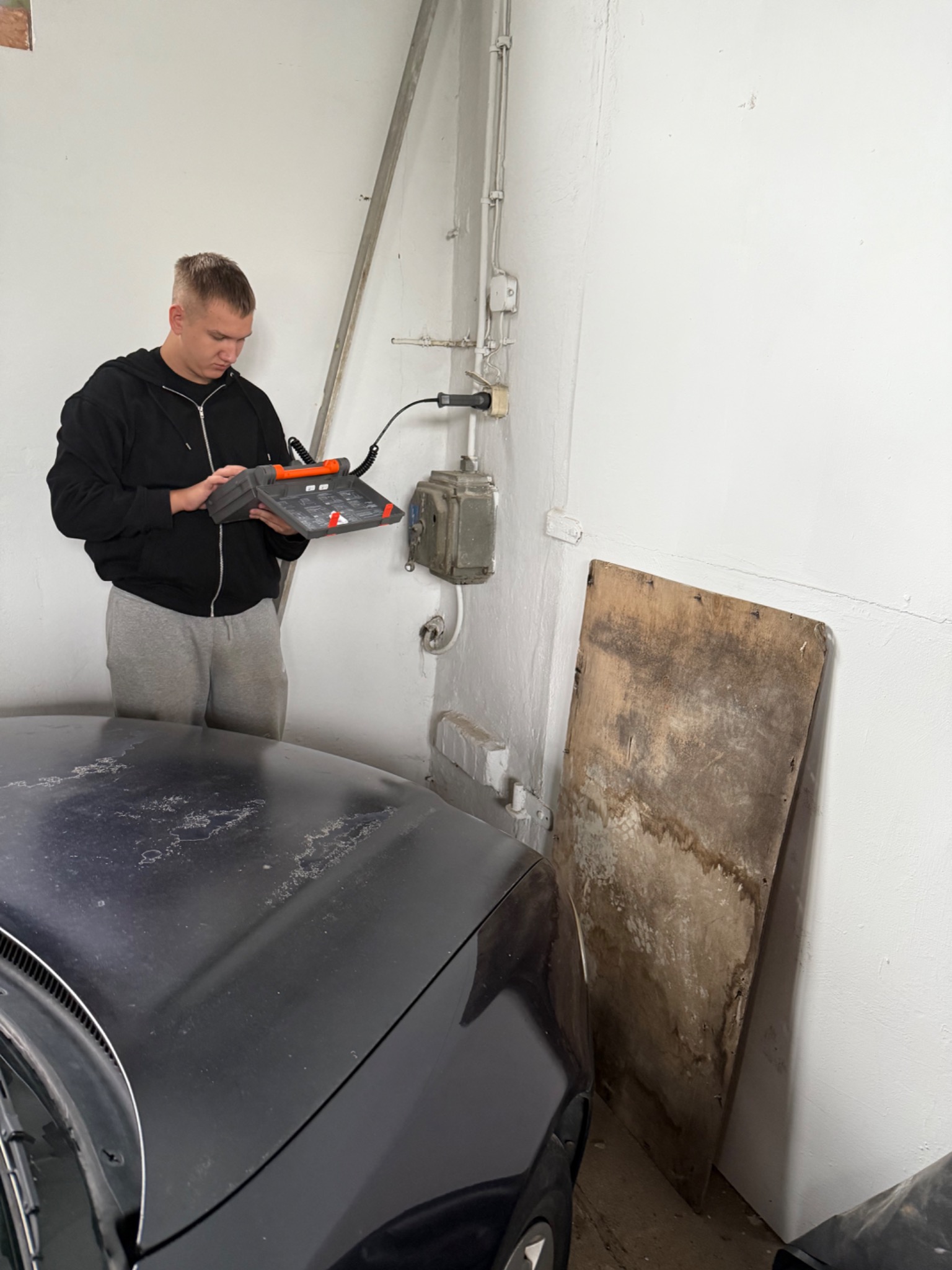 Elektryk sprawdza instalację elektryczną w garażu za pomocą miernika. Widoczne przewody, puszki elektryczne i fragment samochodu.