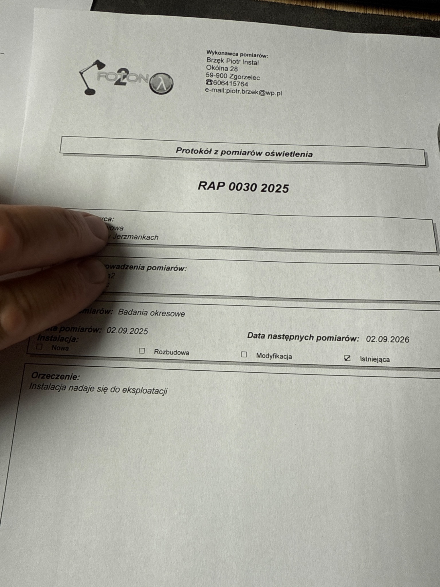 Protokół z pomiarów oświetlenia, dokument z pieczątką firmy Fozon, z datami pomiarów i orzeczeniem o zdatności instalacji do eksploatacji. Częściowo zasłonięty palcem.