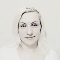 Portret kobiety z blond włosami, skierowanej wprost, z delikatnym uśmiechem na tle jasnej ściany. Zdjęcie w odcieniach szarości, z delikatnym filtrem.