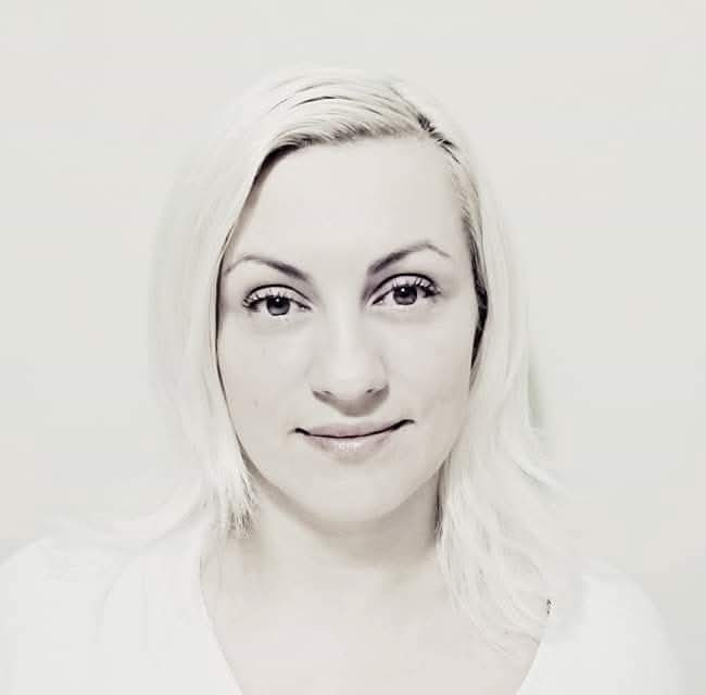 Portret kobiety z blond włosami, skierowanej wprost, z delikatnym uśmiechem na tle jasnej ściany. Zdjęcie w odcieniach szarości, z delikatnym filtrem.