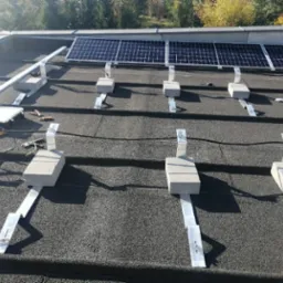 Montaż paneli fotowoltaicznych na dachu pokrytym papą. Widoczne betonowe podstawy i aluminiowe szyny montażowe. Częściowo zainstalowane panele na tle drzew.