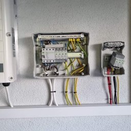 EnergieHelden Natalia Widurska - Instalacja elektryczna: rozdzielnice, inwerter SolarEdge. Widoczne przewody, zabezpieczenia. Montaż na ścianie o nierównej strukturze, listwa przypodłogowa.