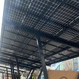 EnergieHelden Natalia Widurska - Montaż paneli fotowoltaicznych na konstrukcji wsporczej. Widoczne panele, stalowe słupy, pracownicy w kamizelkach odblaskowych i karton z logo firmy TommaTech.