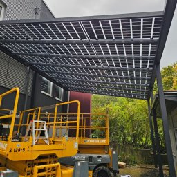 EnergieHelden Natalia Widurska - Montaż paneli fotowoltaicznych na konstrukcji pergoli, widoczny podnośnik koszowy, pochmurne niebo w tle.