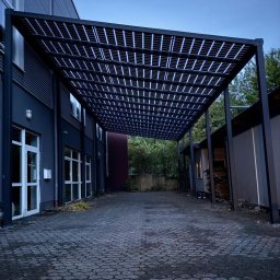 EnergieHelden Natalia Widurska - Nowoczesna, czarna pergola z ażurowym dachem, przylegająca do budynku z dużymi oknami. Kostka brukowa na podłożu, widok na zieleń w tle.