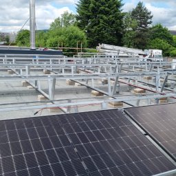 EnergieHelden Natalia Widurska - Montaż paneli słonecznych na dachu budynku w Witkowie. Widoczne panele, metalowa konstrukcja wsporcza i pracownik. W tle zabudowa miejska i zieleń.