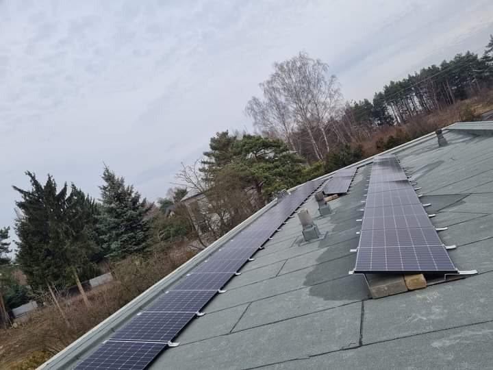 Montaż paneli fotowoltaicznych na dachu pokrytym papą, widok z dołu pod kątem. Panele ułożone w rzędach, w tle drzewa i pochmurne niebo.