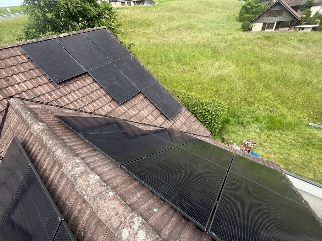 Panele fotowoltaiczne na dachu pokrytym czerwoną dachówką, na tle zielonego wzgórza i domów. Część paneli lekko zabrudzona, widoczne narzędzia i akcesoria montażowe.