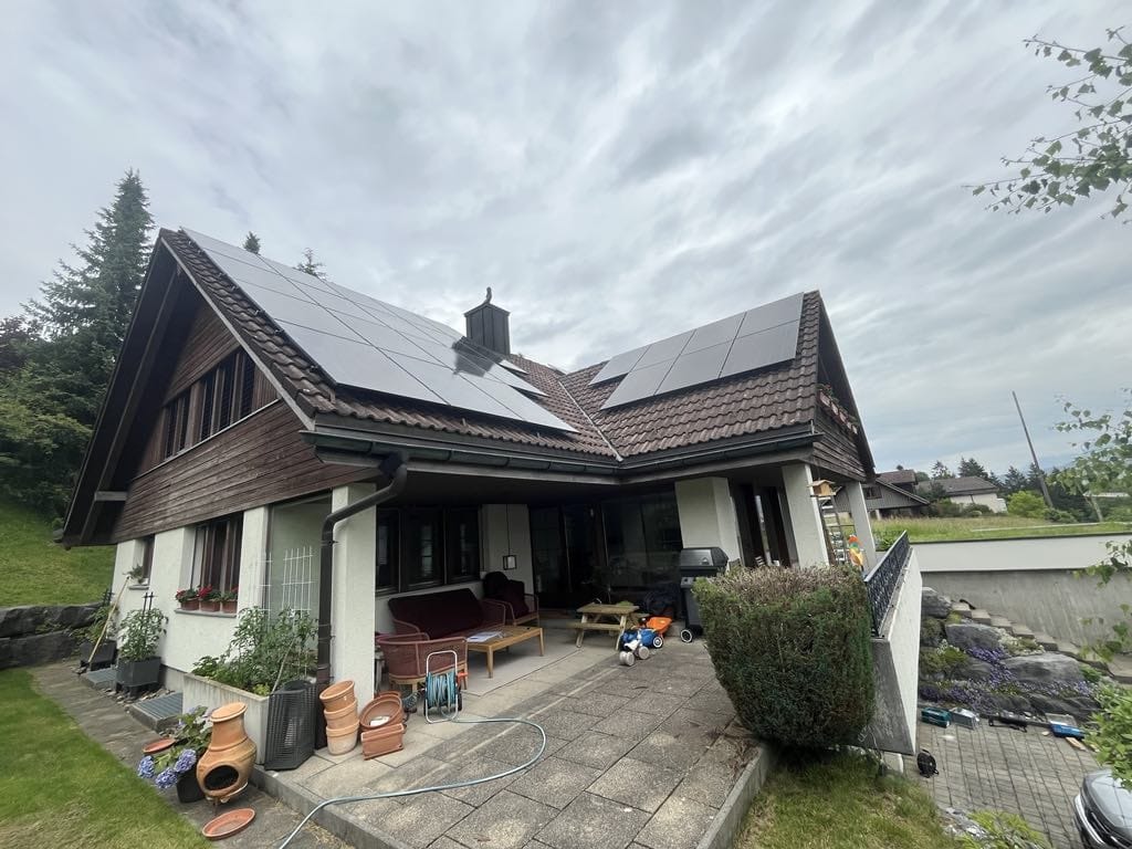 Dom z brązowym dachem i panelami fotowoltaicznymi w słoneczny dzień. Widoczny taras z meblami ogrodowymi i dekoracjami.