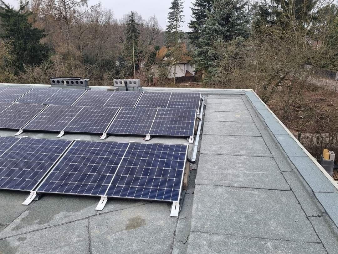 Panele fotowoltaiczne na płaskim dachu z widocznymi elementami montażowymi i kominami wentylacyjnymi w tle drzew.