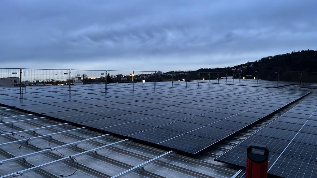 Rozległa instalacja paneli słonecznych na dachu budynku, widoczna konstrukcja montażowa i narzędzie, w tle zabudowa miejska pod pochmurnym niebem.