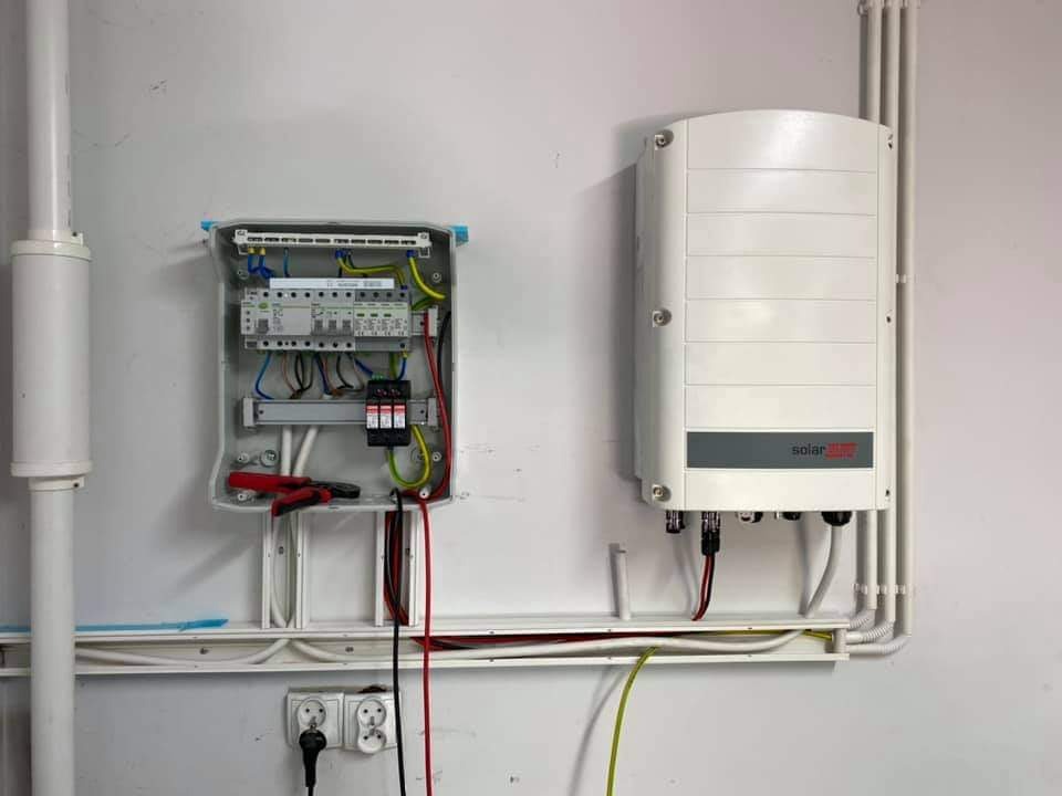 Biały inwerter fotowoltaiczny SolarEdge zamontowany obok otwartej skrzynki elektrycznej z przewodami na białej ścianie. Widoczne gniazdka elektryczne i rury.