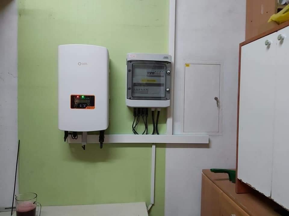 Biały inwerter Solis zamontowany na zielonej ścianie obok skrzynki elektrycznej z licznymi kablami. Widoczny również fragment szafki i szklanka z napojem.