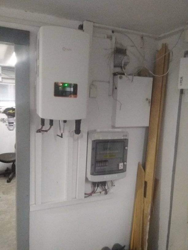 Biały inwerter fotowoltaiczny z zieloną diodą, obok skrzynka elektryczna i licznik na białej ścianie w rogu pomieszczenia. Widoczne kable i rury.