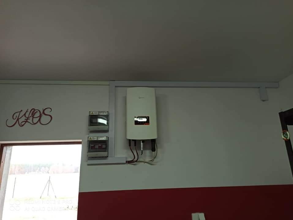 Biały inwerter fotowoltaiczny zamontowany na ścianie, obok dwa liczniki elektryczne. Widoczne przewody i instalacja elektryczna. Na ścianie dekoracyjny napis 'KLOS'.