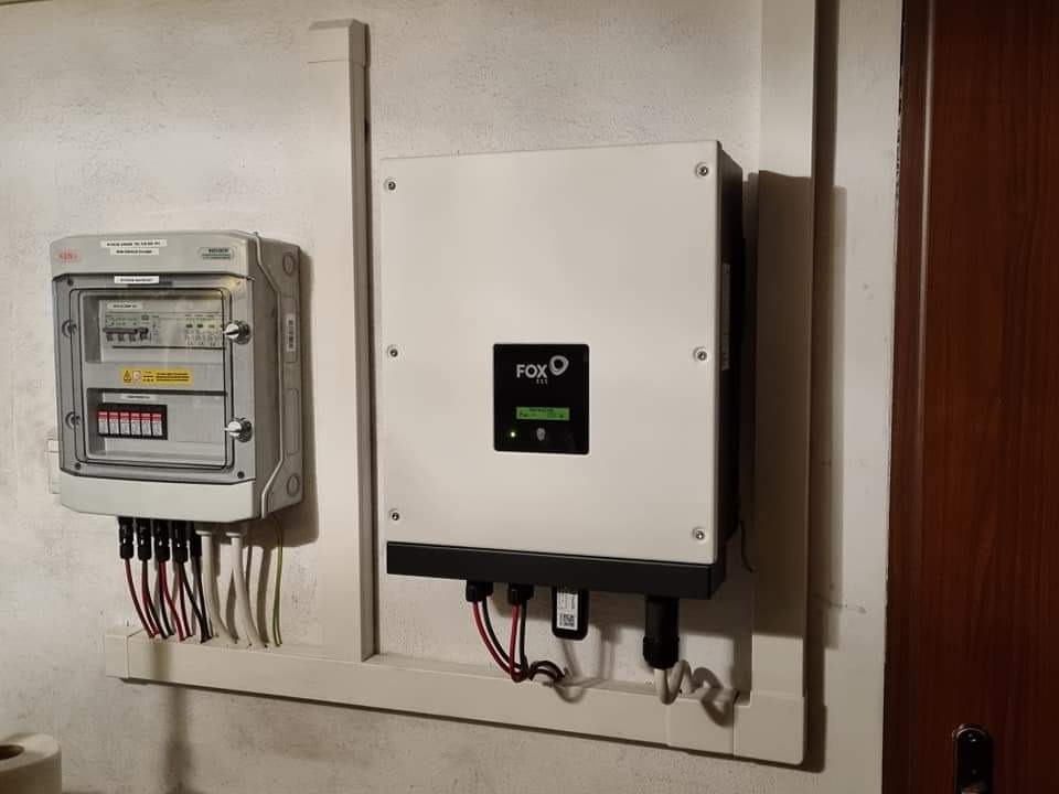 Biały inwerter FOX i skrzynka z bezpiecznikami, połączone przewodami w białych listwach na ścianie. Widoczne złącza kablowe.