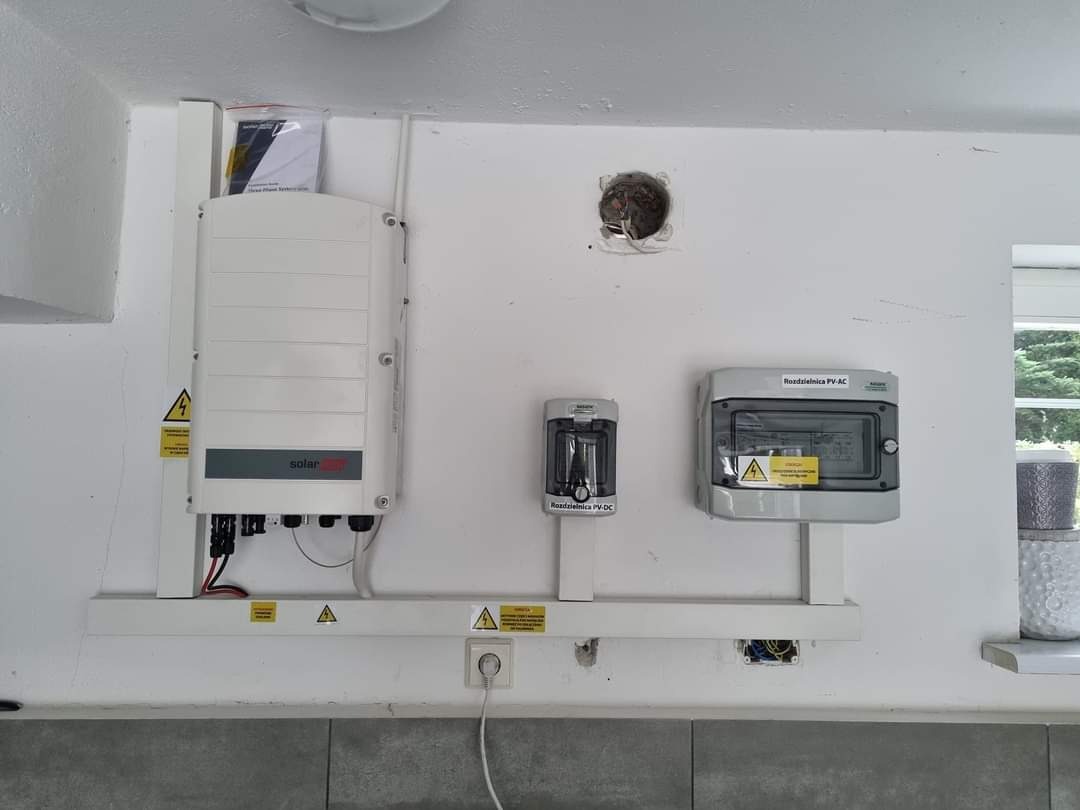 Biała ściana z zamontowanym inwerterem SolarEdge, rozdzielnicą PV-AC i PV-DC. Widoczne kable i oznaczenia ostrzegawcze. Ujęcie z bliska, neutralne oświetlenie.