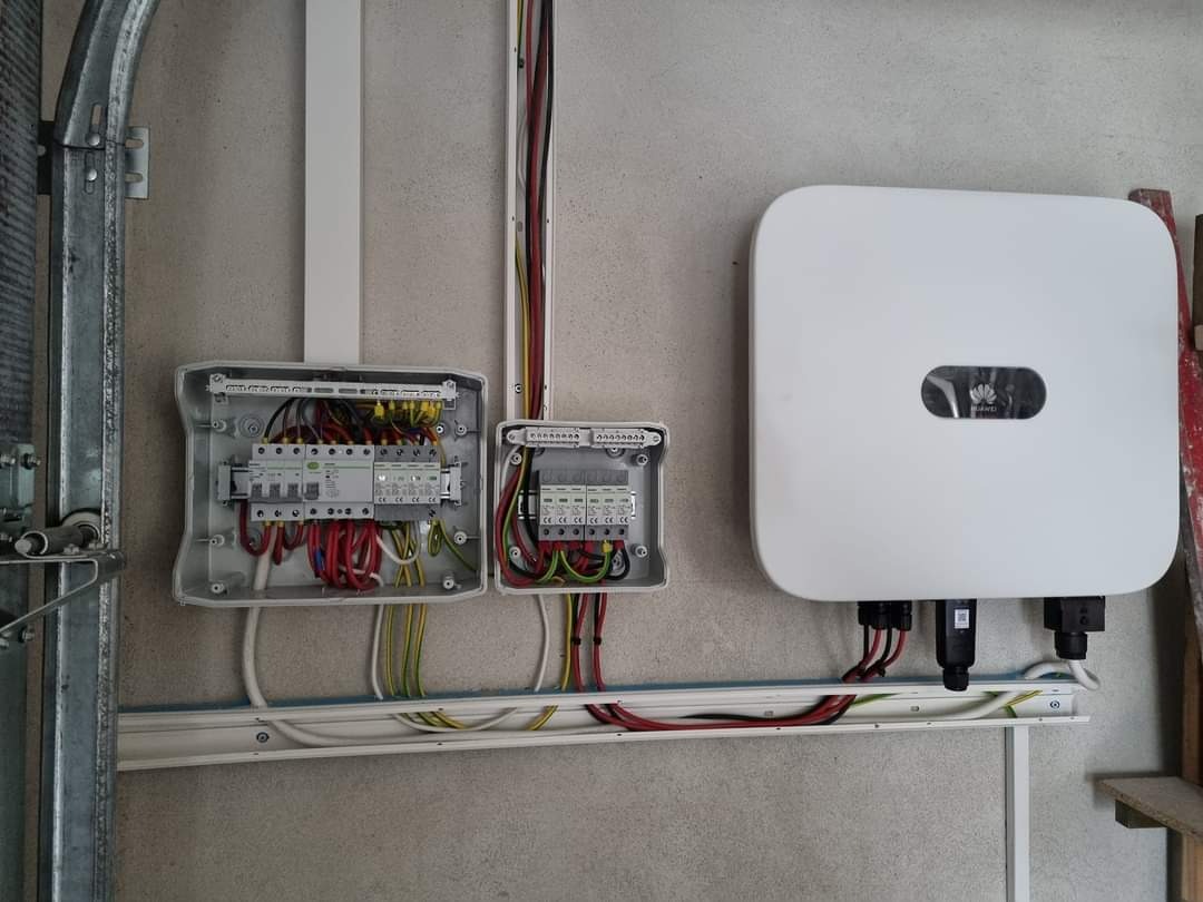 Biały inwerter Huawei i dwie otwarte skrzynki z bezpiecznikami oraz okablowaniem na ścianie. Widoczne przewody i listwy instalacyjne.