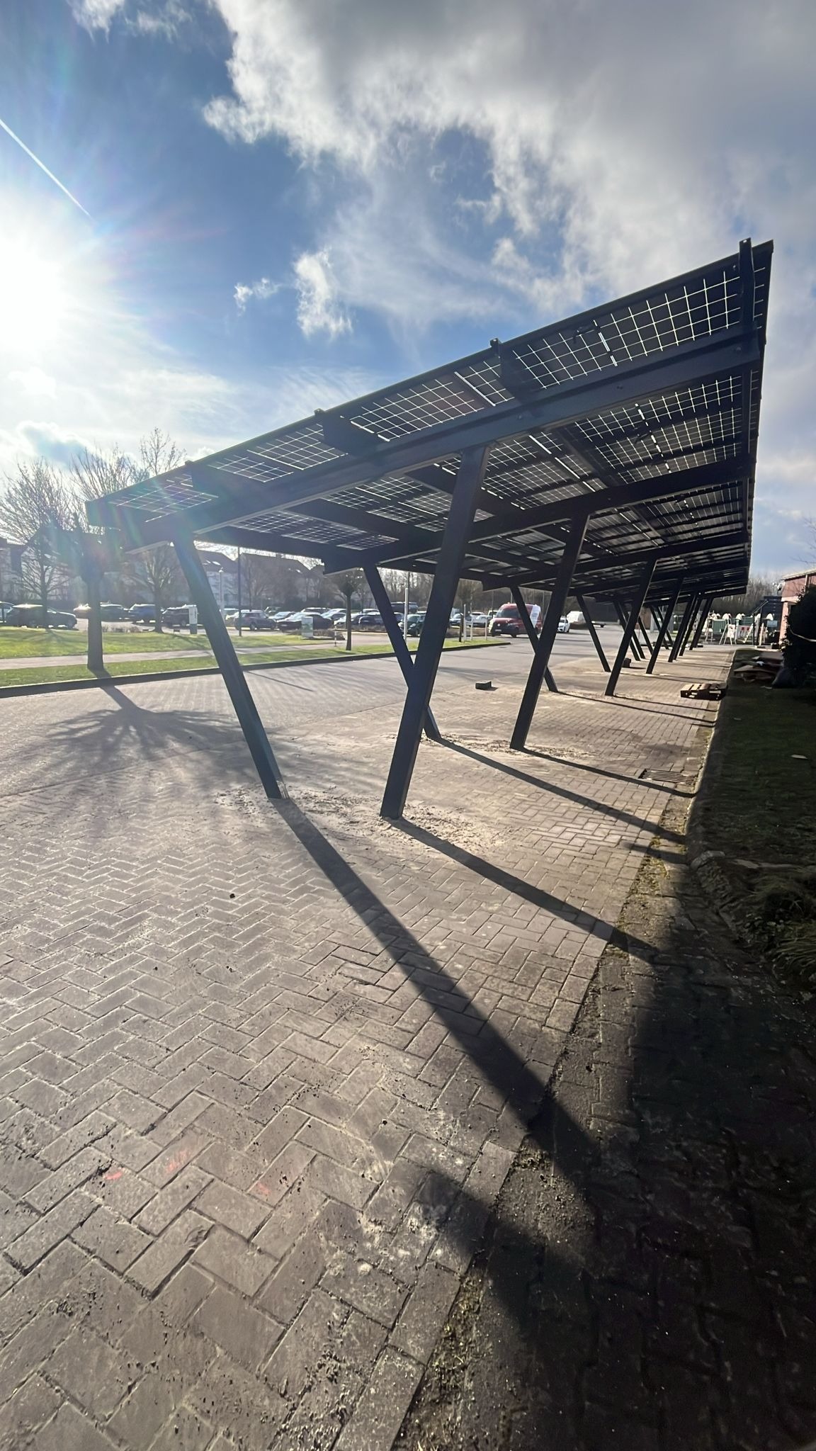 Zadaszenie parkingu z panelami fotowoltaicznymi w słoneczny dzień. Konstrukcja wsparta na ukośnych słupach, rzucających długie cienie na brukowaną nawierzchnię.