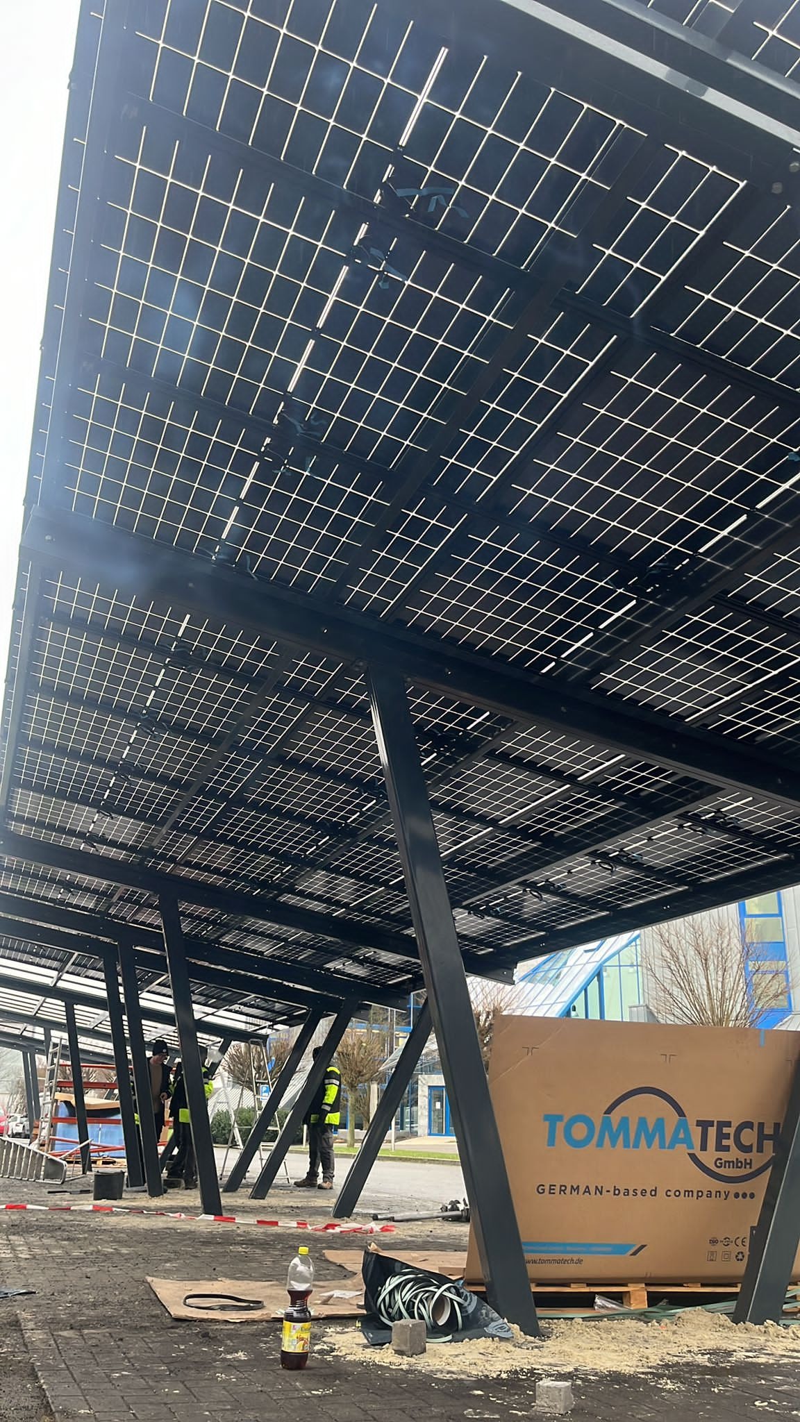 Montaż paneli fotowoltaicznych na konstrukcji wsporczej. Widoczne panele, stalowe słupy, pracownicy w kamizelkach odblaskowych i karton z logo firmy TommaTech.