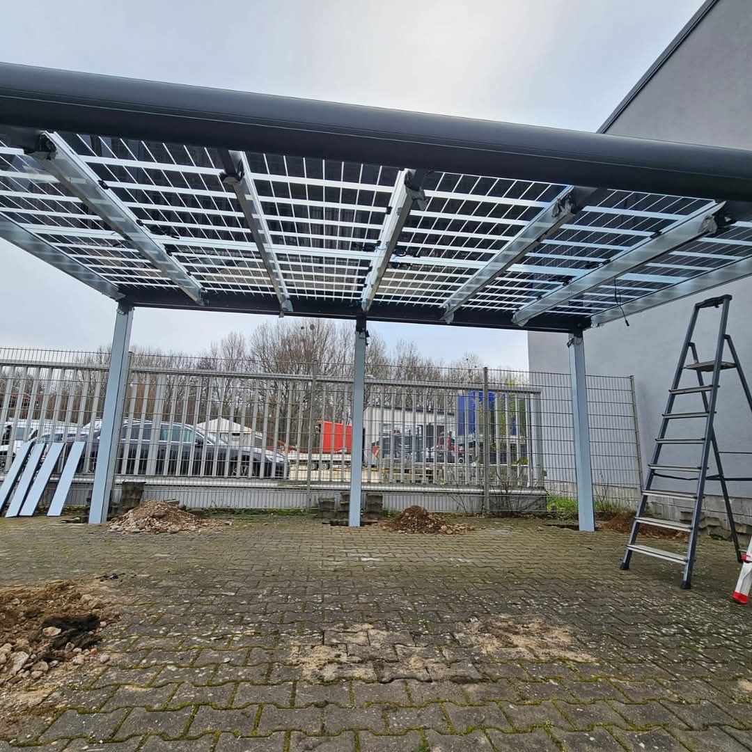 Montaż zadaszenia fotowoltaicznego na parkingu, widoczne panele, metalowa konstrukcja i drabina. Prace w toku, ziemia wokół słupów.