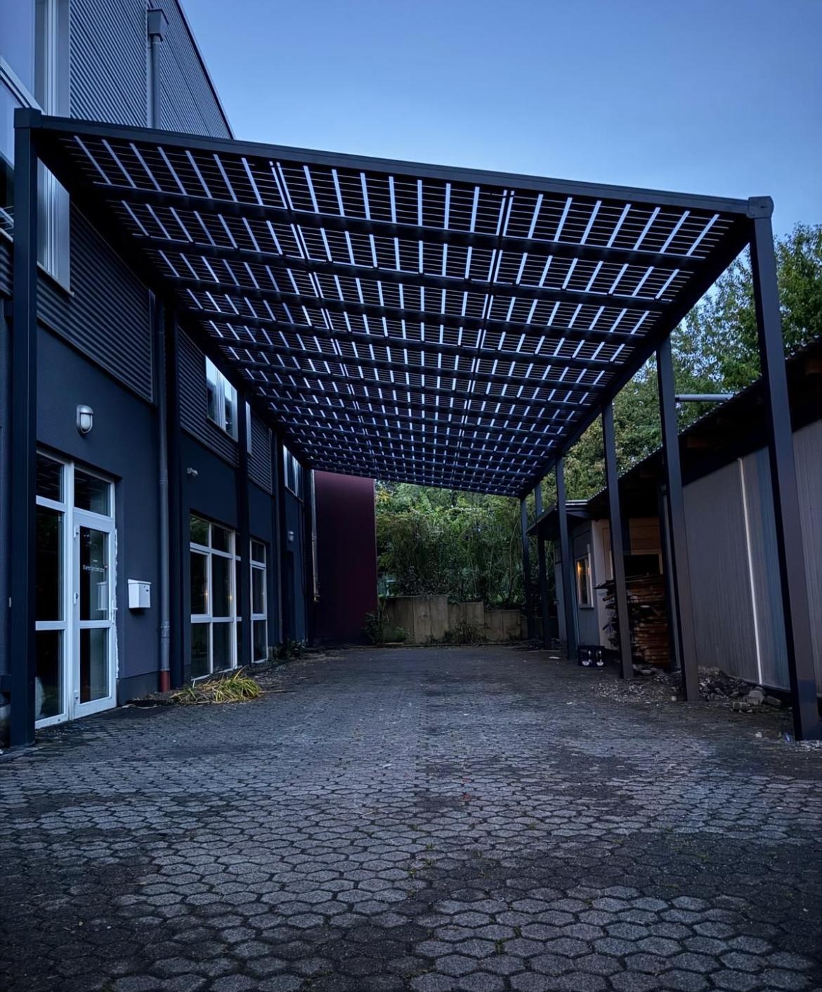 Nowoczesna, czarna pergola z ażurowym dachem, przylegająca do budynku z dużymi oknami. Kostka brukowa na podłożu, widok na zieleń w tle.