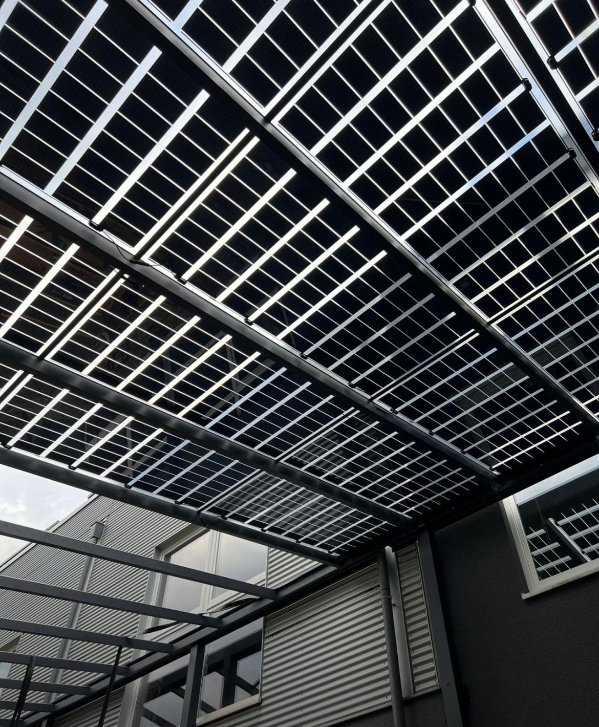 Nietypowy montaż paneli fotowoltaicznych jako zadaszenie nad budynkiem z szarej blachy falistej, widok z dołu pod kątem.