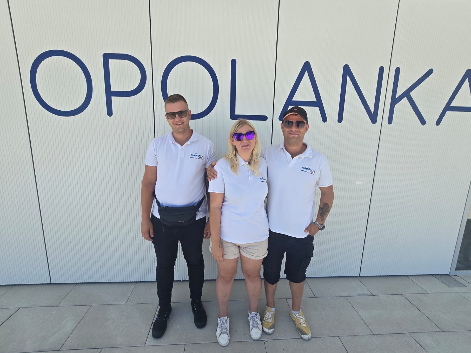 Trzy osoby w białych koszulkach polo z logo firmy na tle białej ściany z napisem 'OPOLANKA'. Mężczyzna z czarną saszetką na biodrach i kobieta w okularach.