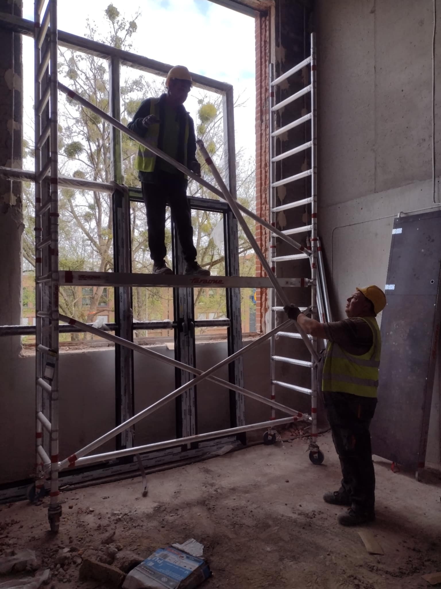 Montaż rusztowania aluminiowego przy oknie przez dwóch pracowników w kaskach i kamizelkach odblaskowych. W tle widoczne drzewa.