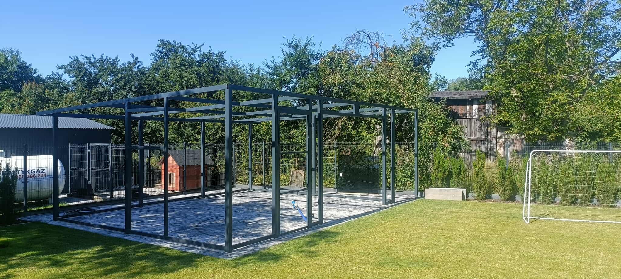 Konstrukcja metalowej pergoli na posesji, widok z zewnątrz. Szara, geometryczna rama na kostce brukowej, zieleń drzew w tle, część otoczenia.