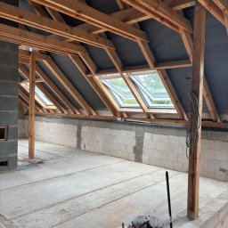 RENOVIO Sp. z o.o. - Poddasze w stanie surowym otwartym: drewniana konstrukcja dachu z oknami połaciowymi, ściany z białych bloczków, betonowa wylewka. Widoczne narzędzia i materiały budowlane.