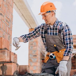 RENOVIO Sp. z o.o. - Murarz w kasku i okularach ochronnych, w trakcie murowania ściany z czerwonej cegły z użyciem kielni. Widoczne narzędzia w pasie.