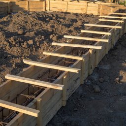 RENOVIO Sp. z o.o. - Szczegółowe ujęcie drewnianego szalunku zbrojonego stalą, przygotowanego do wylania fundamentów pod budowę domu. Widoczne elementy konstrukcji i ziemia.