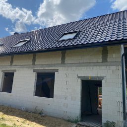 RENOVIO Sp. z o.o. - Niedokończony dom z białego bloczka, widok z zewnątrz. Widać otwory okienne i drzwiowe, czarny dach z oknami dachowymi i fragment nieba z chmurami.