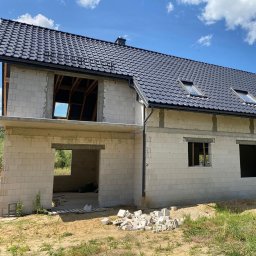 RENOVIO Sp. z o.o. - Dom w stanie surowym otwartym z czarną dachówką i oknami dachowymi. Widoczne białe bloczki i niezadaszony otwór wejściowy. Częściowo ułożone bloczki na ziemi.